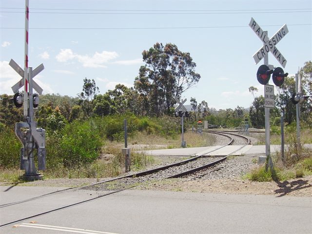 The view looking west into the Newstan Colliery site. The main line at Fassifern is approx. 200 metres behind the camera. The level crossing with boom gates in the foreground is a public road, and the adjacent crossing (lights but no booms) is on an private internal road for coal trucks. In the distance the track on the right is the northern leg of the triangle junction with the main line.