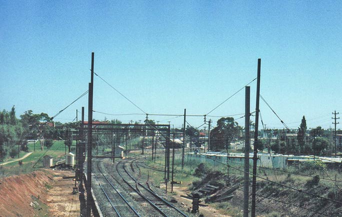 The view looking south towards Regents Park from the former Jenkins St. pedestrian bridge.  Inbetween the track can be seen the Regents Park Signal Box. The one-time branch to Potts Hill reservoir left the main line on the left side at this point. The Commonwealth Siding can be seen leading behind the bank on the right.